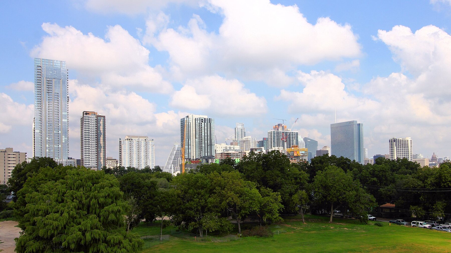 High-rise towers of downtown Austin under a blue sky with white clouds, seen across a park lawn and a line of green trees