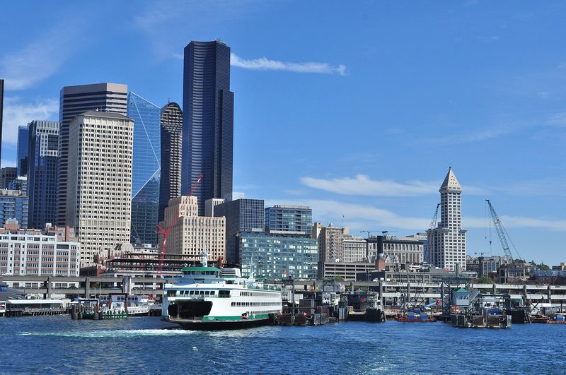 Downtown Seattle skyscrapers under a blue sky, seen from the water of Elliott Bay, with a green and white ferry at the waterfront dock