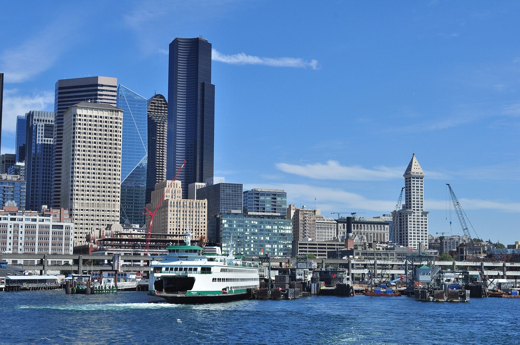Downtown Seattle skyscrapers under a blue sky, seen from the water of Elliott Bay, with a green and white ferry at the waterfront dock