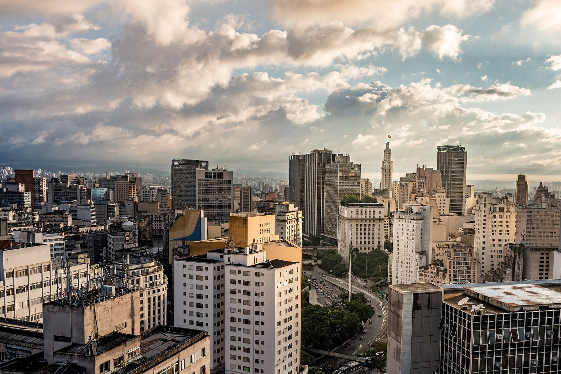High-rise apartment and office towers in central São Paulo under a wide sky of broken cloud, with a curving avenue and trees between the buildings and one tower topped by a flagpole on the skyline.