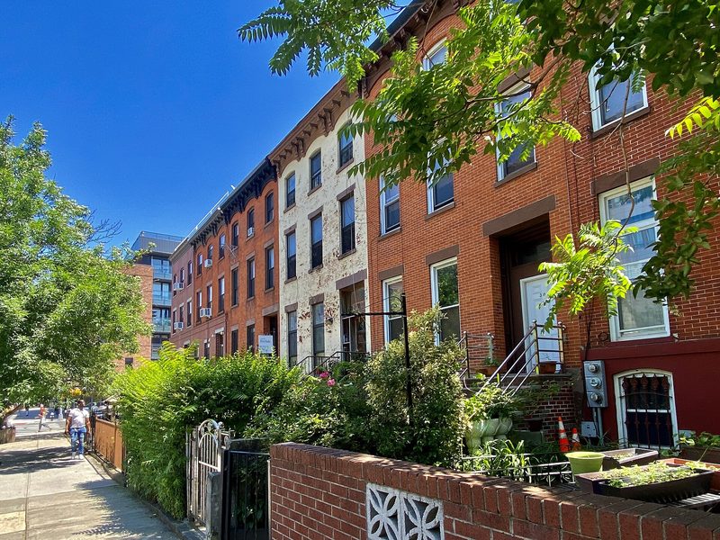 Red-brick and white-painted row houses with stoops and small front gardens line a sunny street in Brooklyn under a blue sky, with a newer apartment block at the far end.
