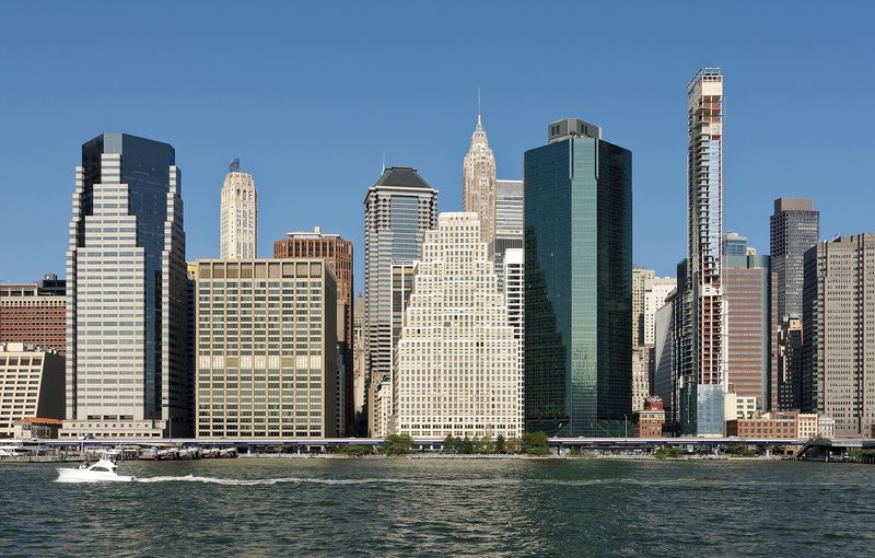 Glass and stone office towers line the Manhattan waterfront under a clear blue sky, seen across the East River, with a small white motorboat crossing in the foreground.