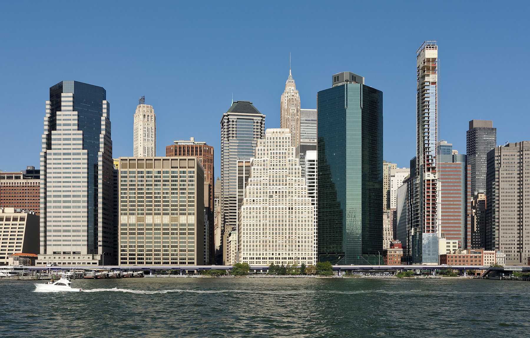 Glass and stone office towers line the Manhattan waterfront under a clear blue sky, seen across the East River, with a small white motorboat crossing in the foreground.