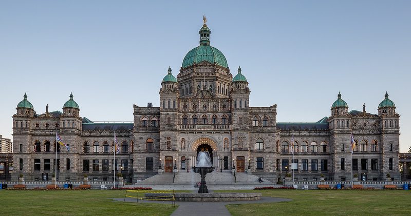 A wide grey stone legislature building with green copper domes and a gilded statue on the central dome, seen across a lawn with a fountain and flagpoles under a clear evening sky.