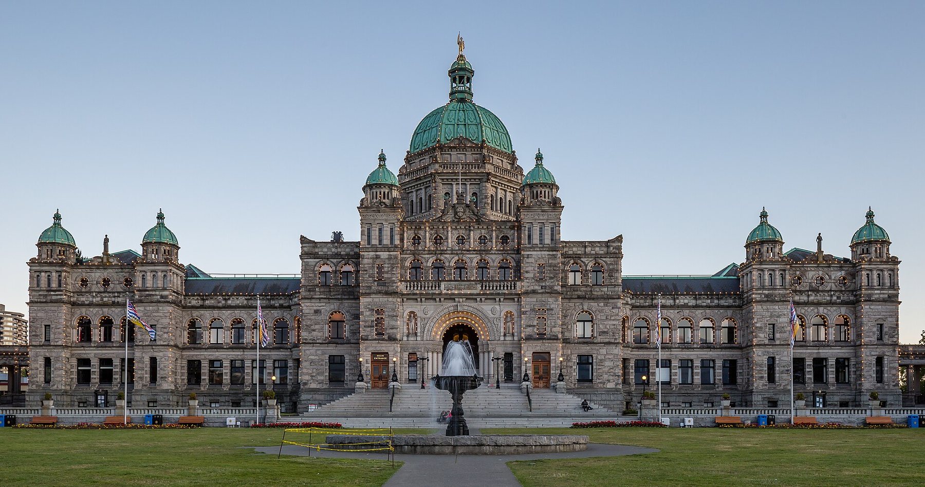 A wide grey stone legislature building with green copper domes and a gilded statue on the central dome, seen across a lawn with a fountain and flagpoles under a clear evening sky.