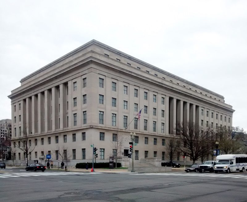 A large pale limestone government building with tall columned bays and an American flag over the corner entrance, seen across a street intersection with cars and a shuttle bus on an overcast day.