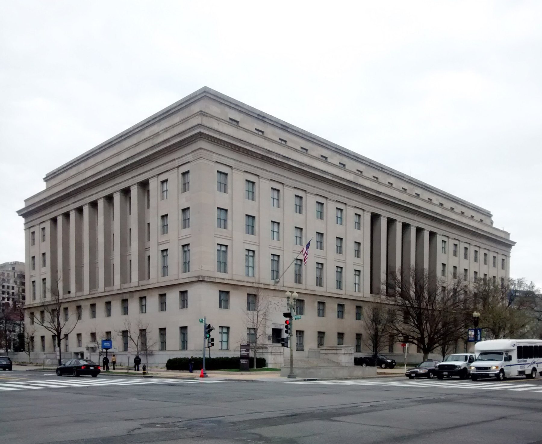A large pale limestone government building with tall columned bays and an American flag over the corner entrance, seen across a street intersection with cars and a shuttle bus on an overcast day.