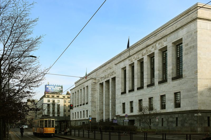 The pale stone front of the Palace of Justice in Milan under a blue sky, with a yellow tram passing on the street below.