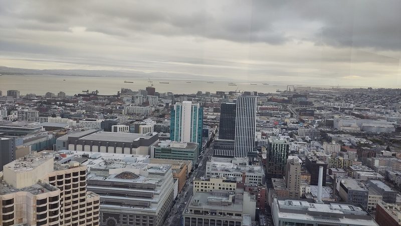 A wide view over San Francisco from a high floor under grey cloud, with towers in the foreground, low city blocks behind them and the bay in the distance.