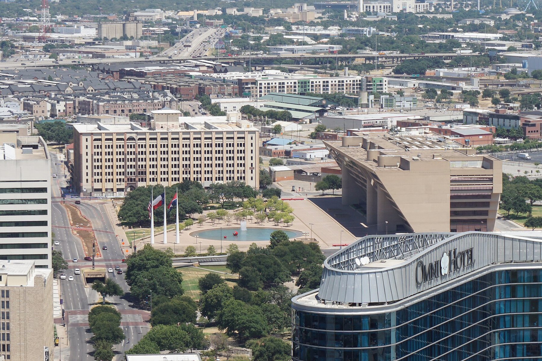 Dallas City Hall, a sloping concrete building, seen from above with its plaza, round reflecting pool and three flagpoles, and the glass roof of a hotel in the foreground.