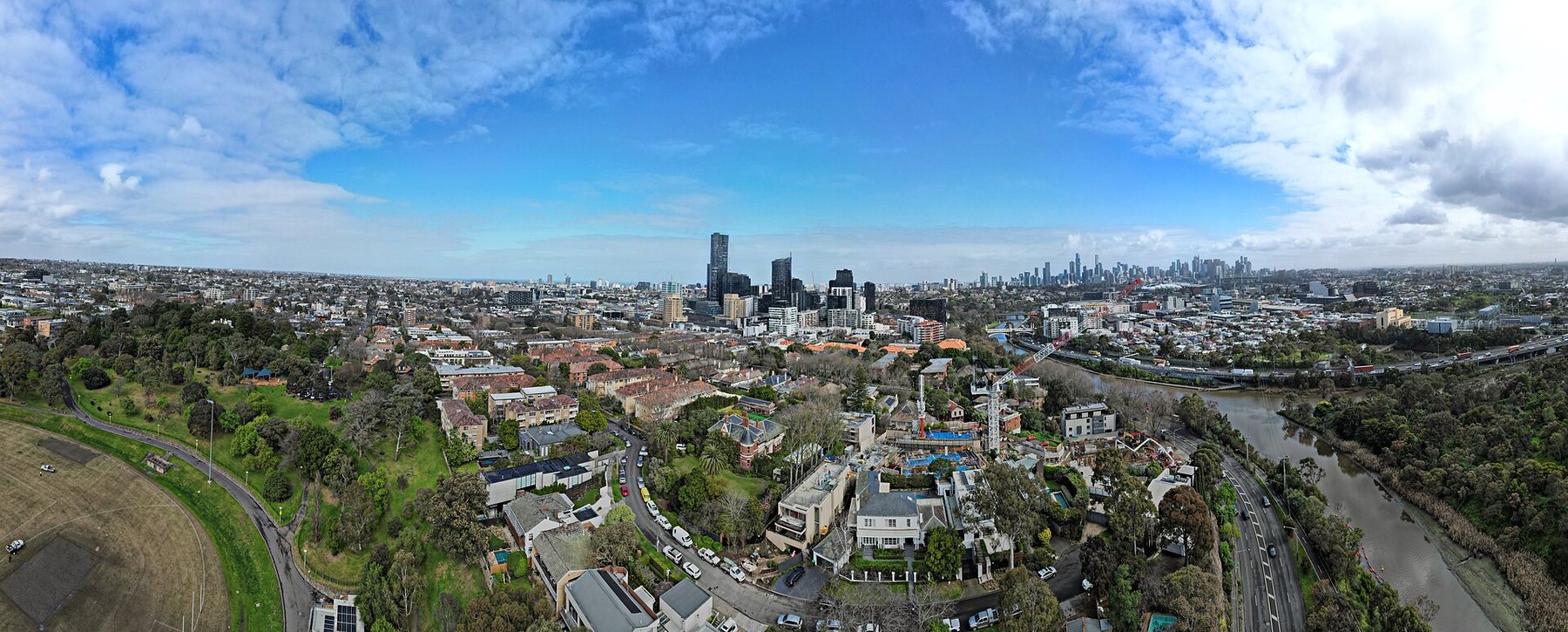 A wide aerial view over houses and tree-lined streets in South Yarra, with a river on the right and the towers of central Melbourne on the horizon under a blue sky with clouds.