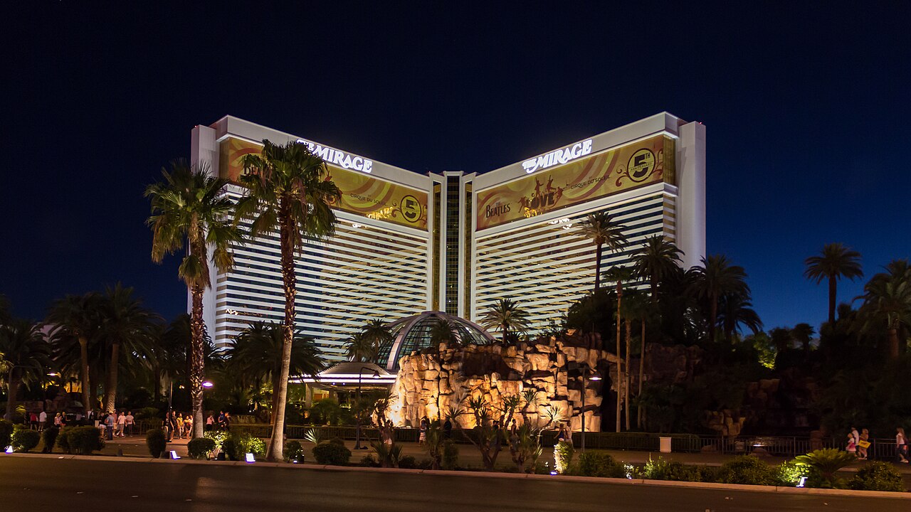 A wide white hotel with three wings and gold-lit bands, behind palm trees under a deep blue evening sky.