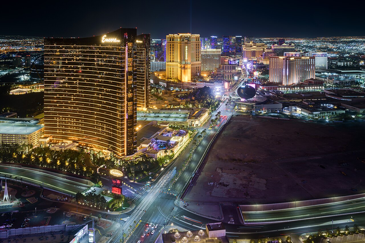 A city of lit hotel towers and bright boulevards at night seen from high above, with a gold glass tower on the left.
