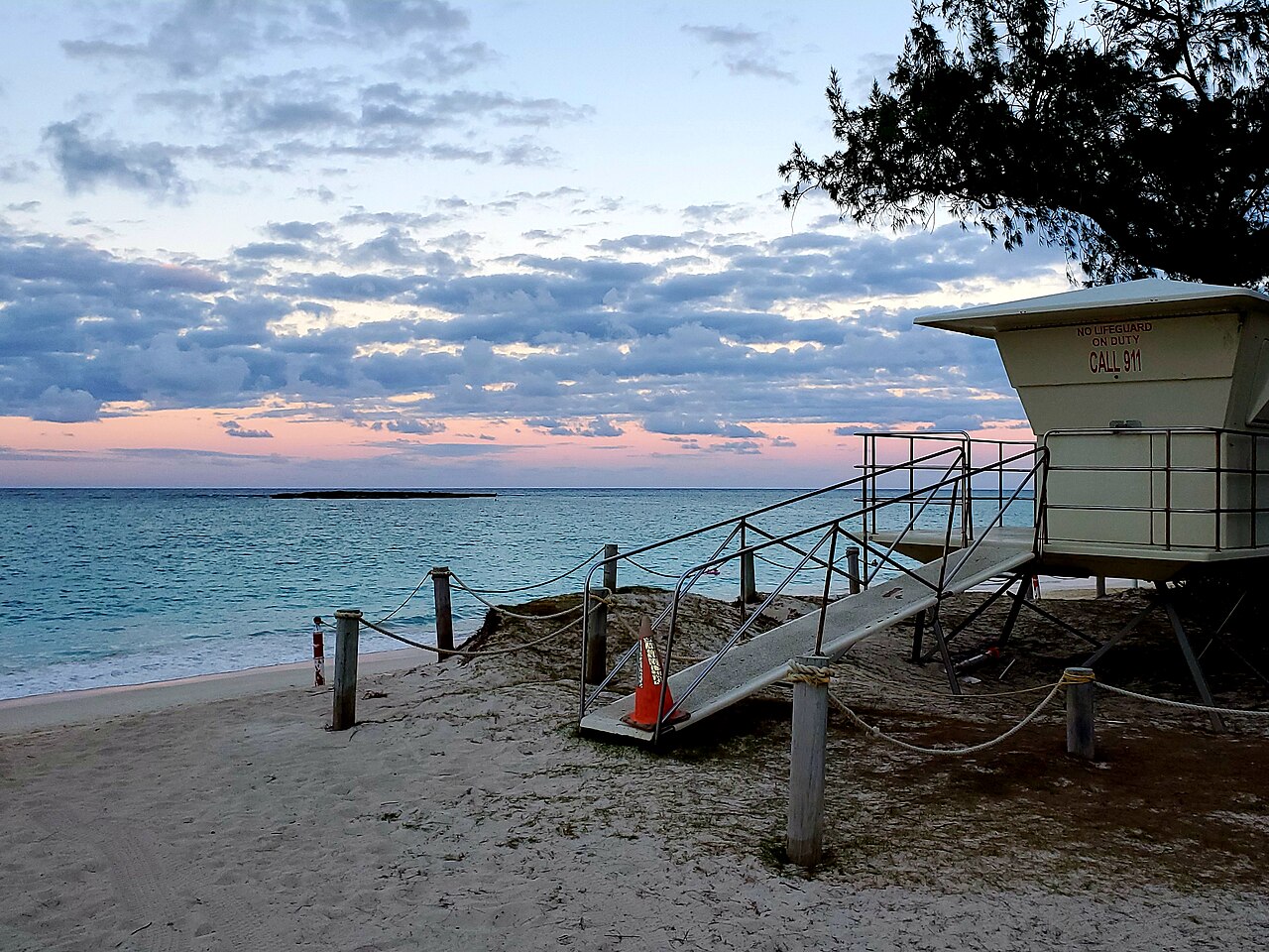 A white lifeguard tower with a ramp on a sandy beach, calm blue water behind it and pink clouds low in the sky.