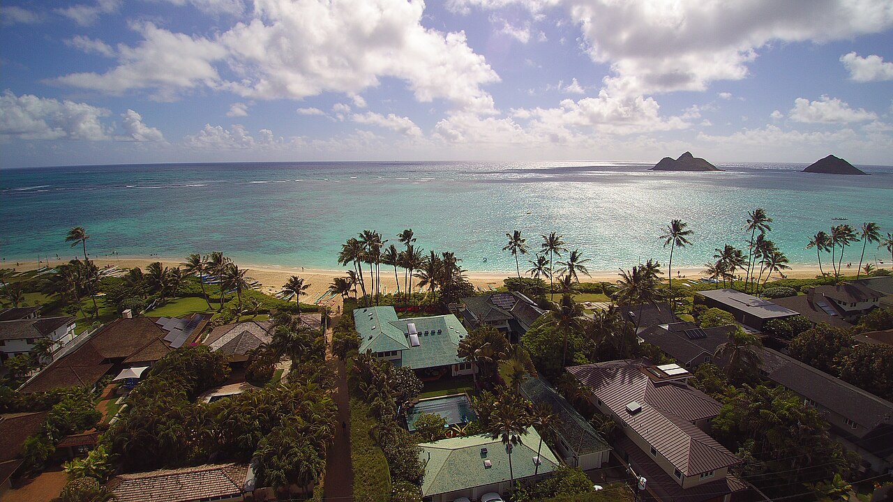 Rooftops and palm trees running down to a pale turquoise bay, with two small islands on the horizon.