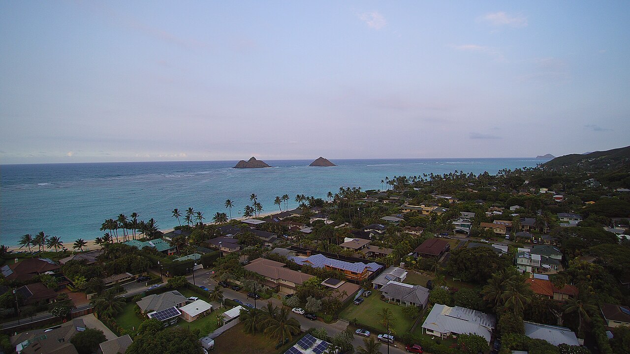 A neighbourhood of low houses and trees beside the sea seen from above at dusk, with two small islands offshore.