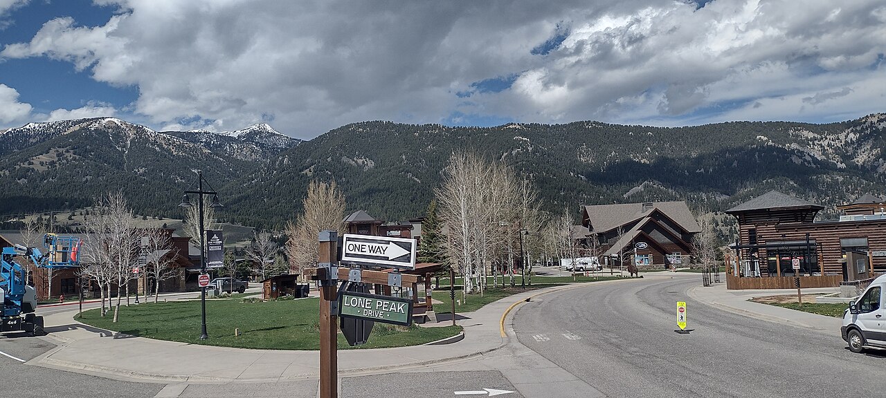 A curving road past timber lodge-style buildings and bare trees, with forested mountains and heavy cloud behind.