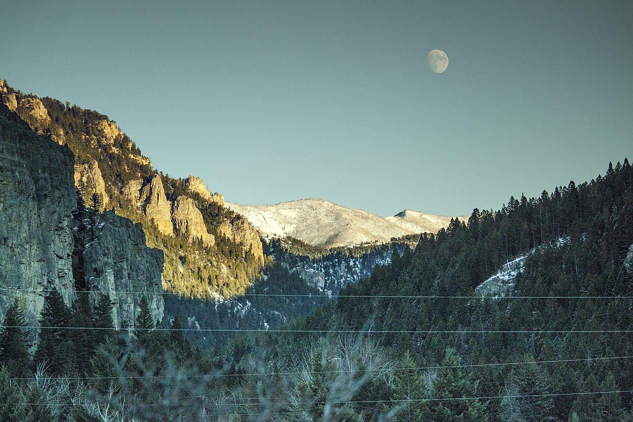 A forested valley in shadow with sunlit cliffs on the left, a snowy ridge in the distance and the moon in a clear sky.