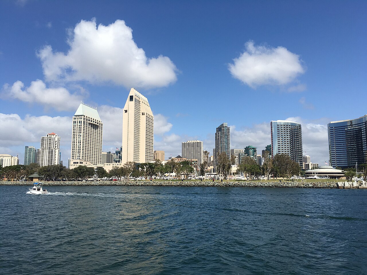 Hotel and apartment towers lined up along a waterfront, seen across dark blue water under a sky with white clouds.