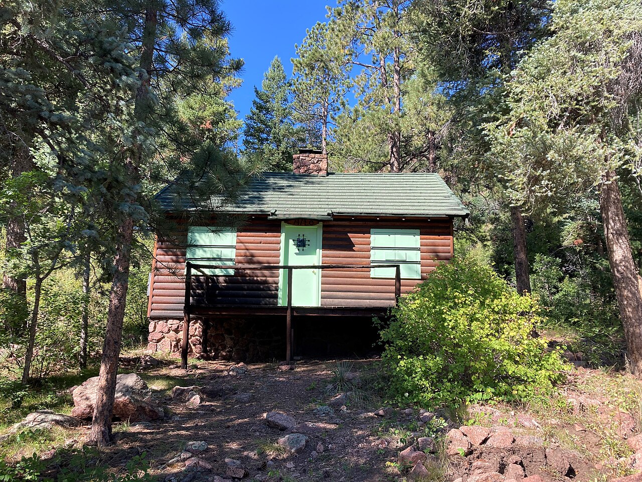 A small log cabin with green shutters and a small wooden porch nestled among evergreen trees.