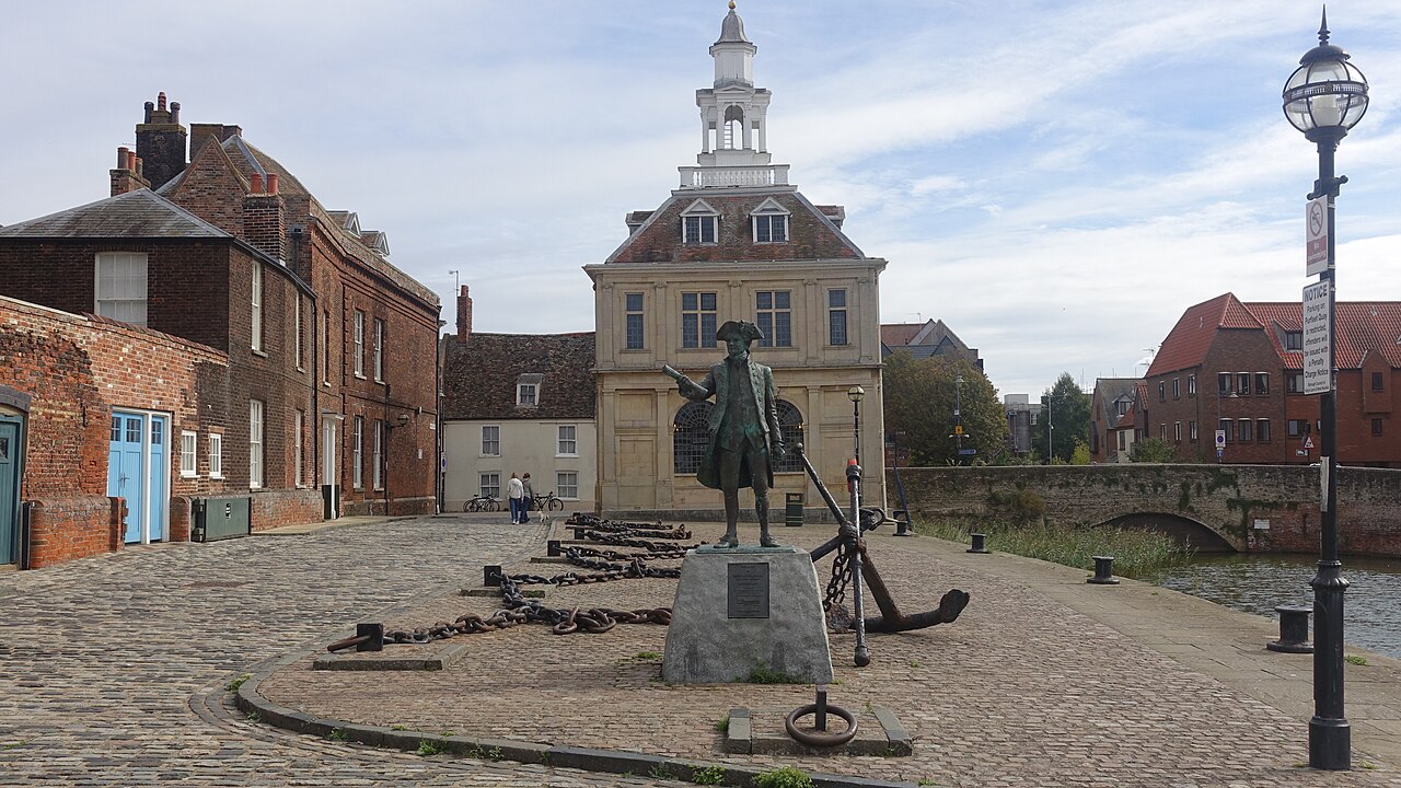 A small square stone building topped by a white lantern tower beside a cobbled quay, with a statue and old brick houses nearby.