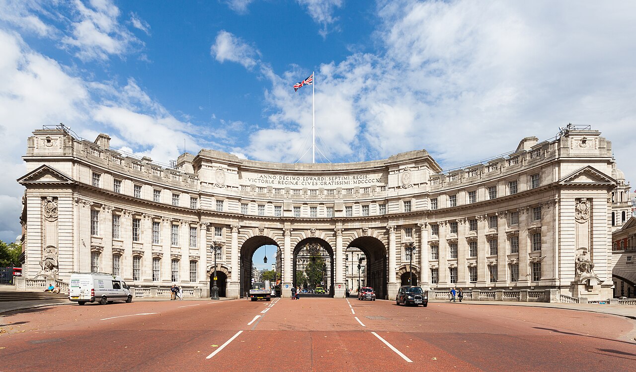 A curved pale stone building with three large arched gateways over a red road, a flag flying above it under a blue sky.