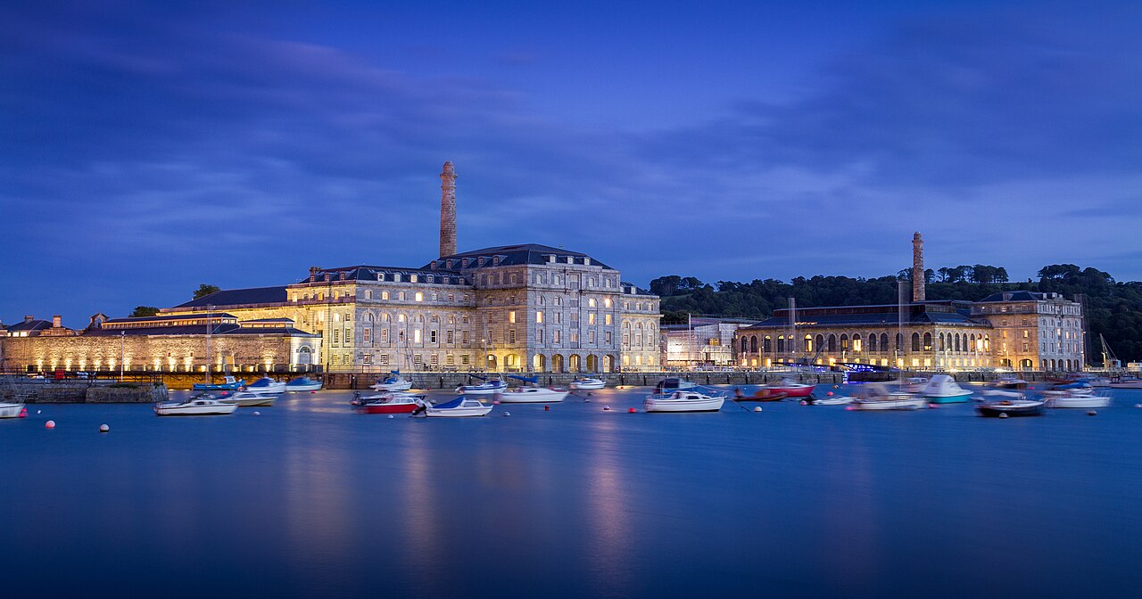 Long floodlit stone buildings with a tall chimney on a waterfront, small boats moored in still blue water in front.