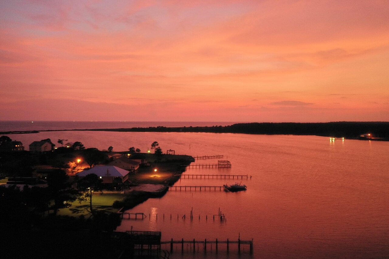 A dark shoreline with piers reaching into flat water that glows orange and pink under a red evening sky.