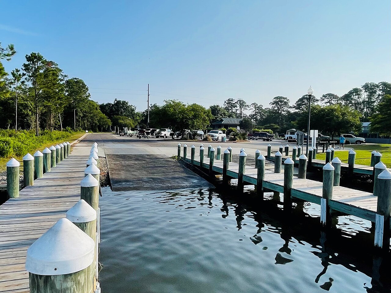 A wooden boardwalk with white-capped posts beside a calm boat ramp, with trees and clear blue sky beyond.