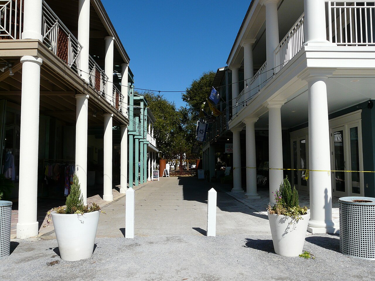 A paved lane between white two-storey buildings with tall columns and balconies, with planters and bollards along it.