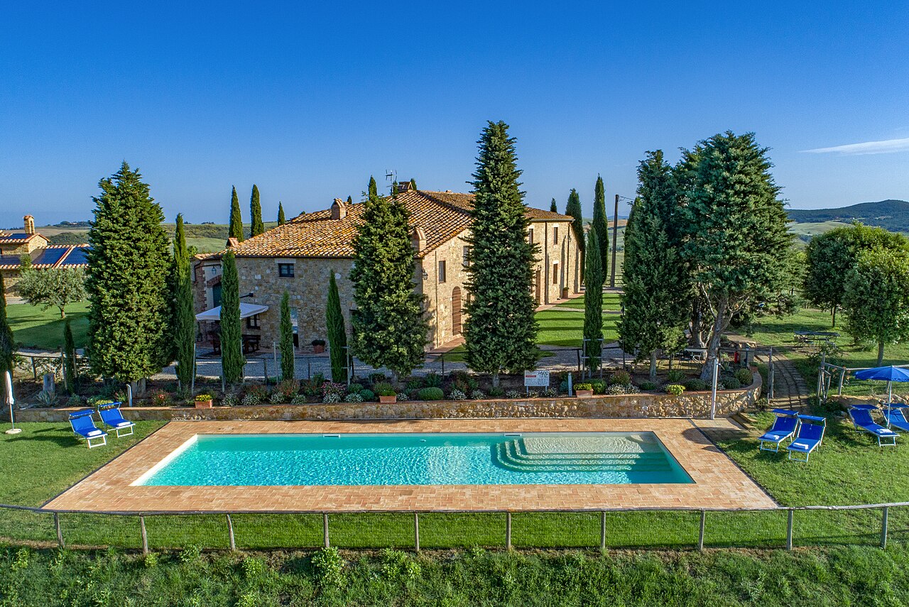 A stone farmhouse among tall cypress trees above a swimming pool and lawn, with rolling green hills behind.