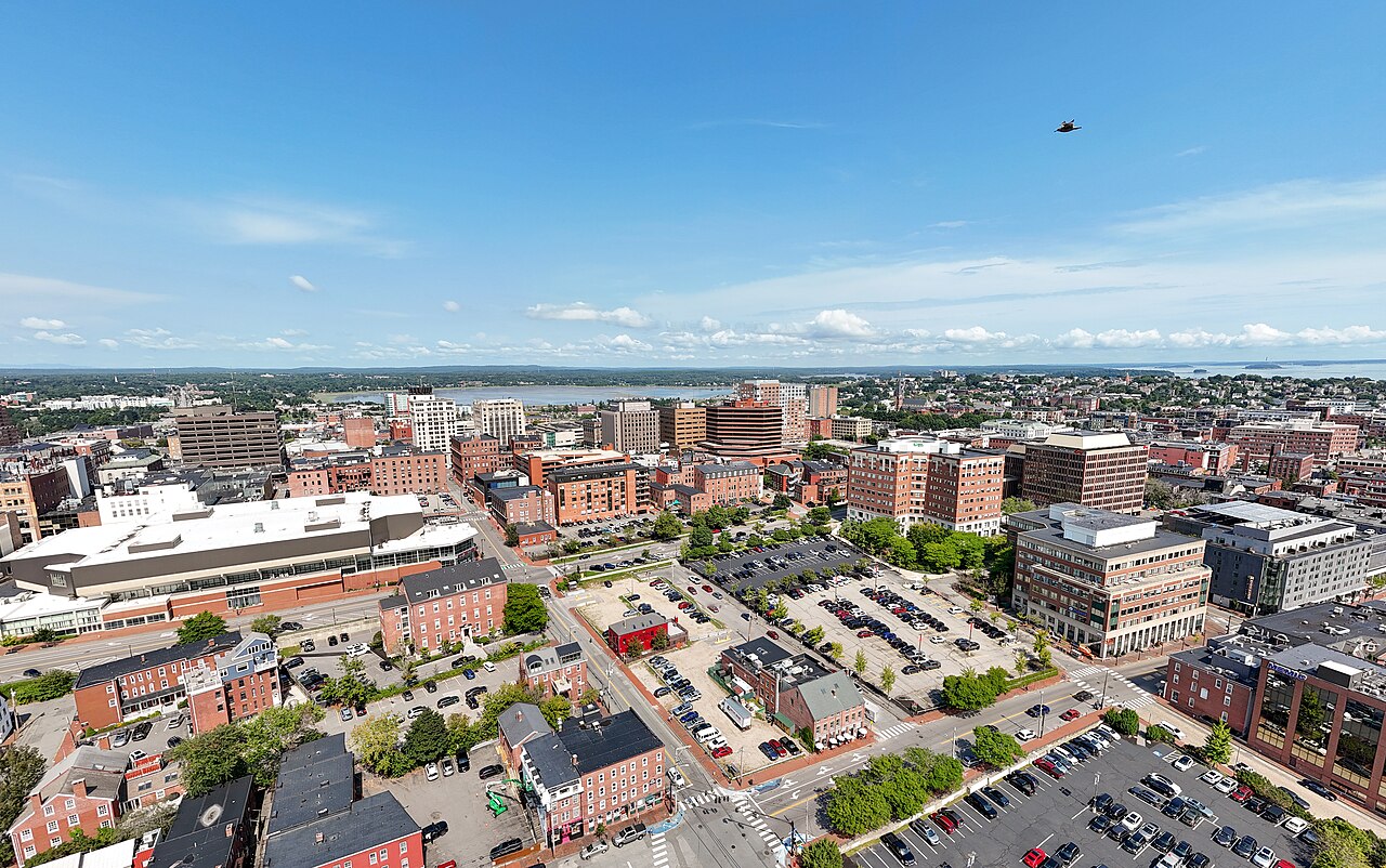 A compact city of red-brick blocks and car parks seen from above, with water and low islands on the horizon.
