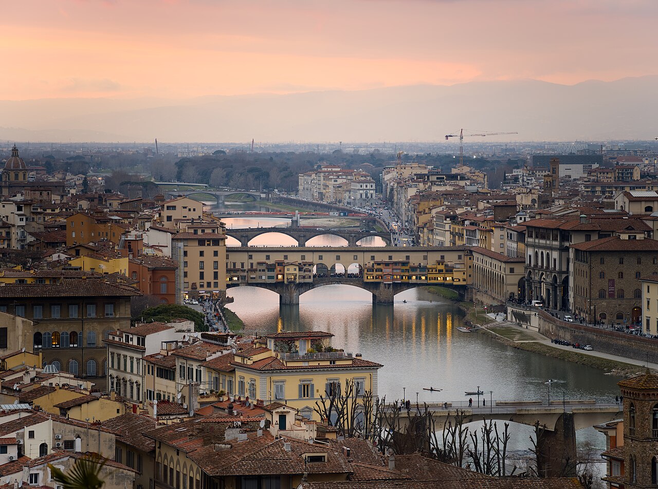 An old bridge lined with small buildings crossing a river between terracotta rooftops, under a hazy pink sky.