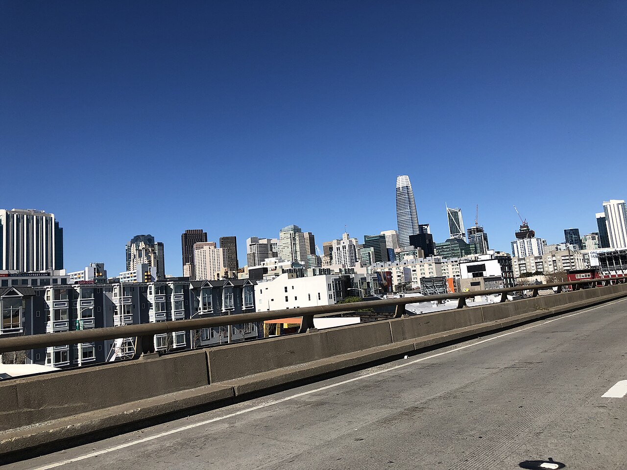 A row of downtown towers against a deep blue sky, seen over the concrete barrier of an elevated road.