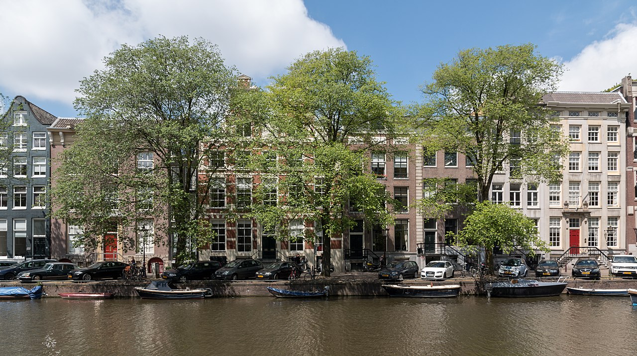 A row of tall narrow brick houses behind leafy trees, with small boats moored along the canal in front.