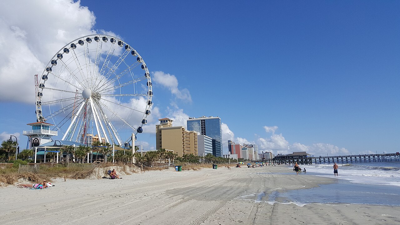 A large white observation wheel stands beside a wide sandy beach with hotel towers and a pier behind it.