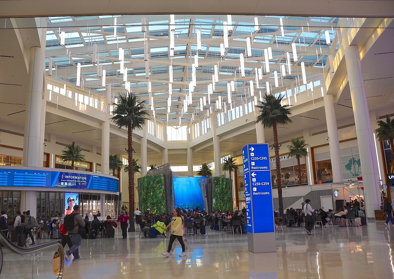 A bright airport hall with a glass roof, tall palm trees and travellers crossing a polished floor.