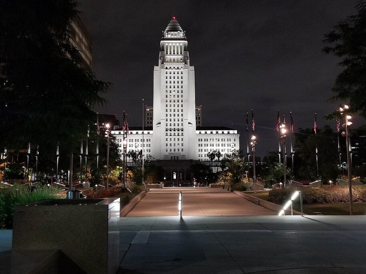 A floodlit white tower with a stepped pyramid top rises above a dark park path lined with flagpoles.