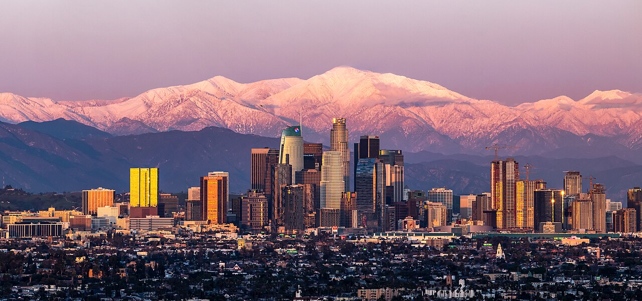 A cluster of skyscrapers glows orange in low sun in front of pink snow-covered mountains.