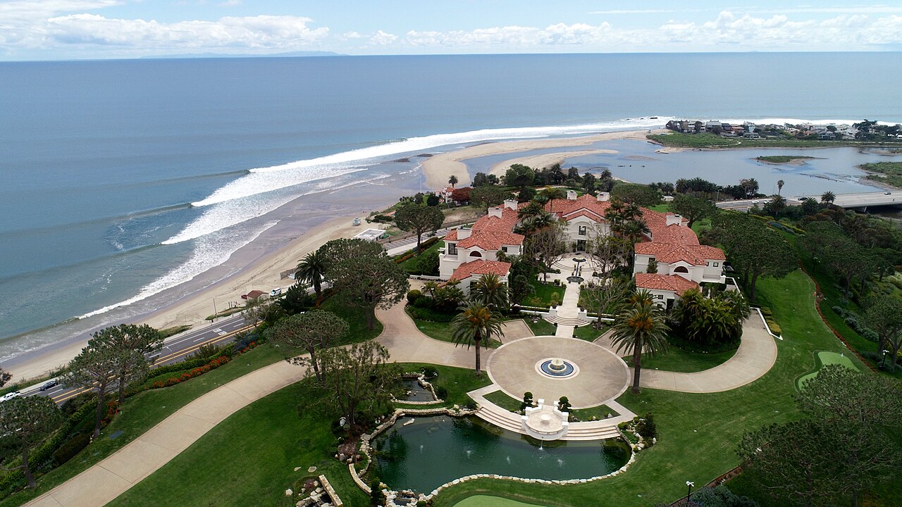 An aerial view of a red-roofed estate with formal gardens beside a curving sandy beach and lines of breaking waves.