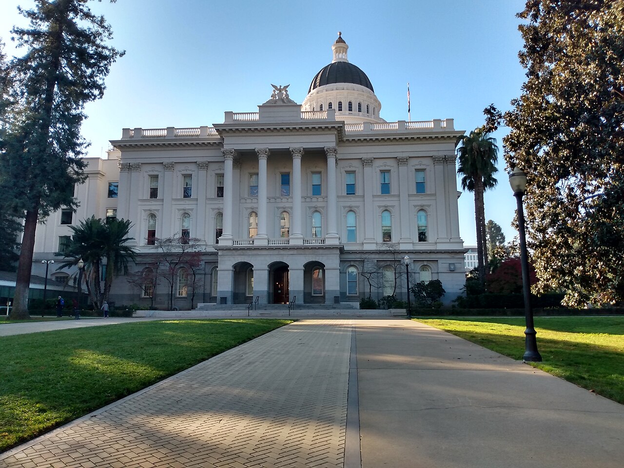 A white neoclassical building with columns and a dome stands at the end of a wide path between lawns and trees.