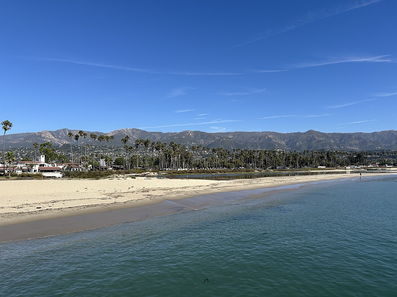 Calm blue water meets a long sandy beach lined with palms, with a mountain ridge behind the town.