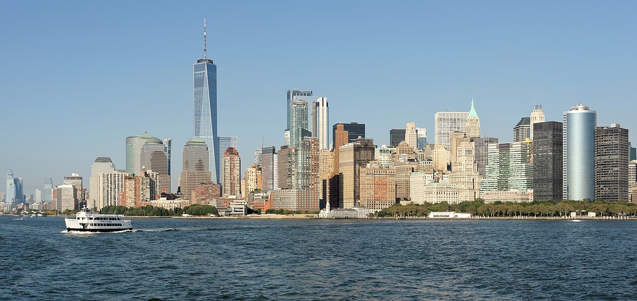 A dense skyline of glass and stone towers rises straight from the edge of a wide stretch of harbour water.