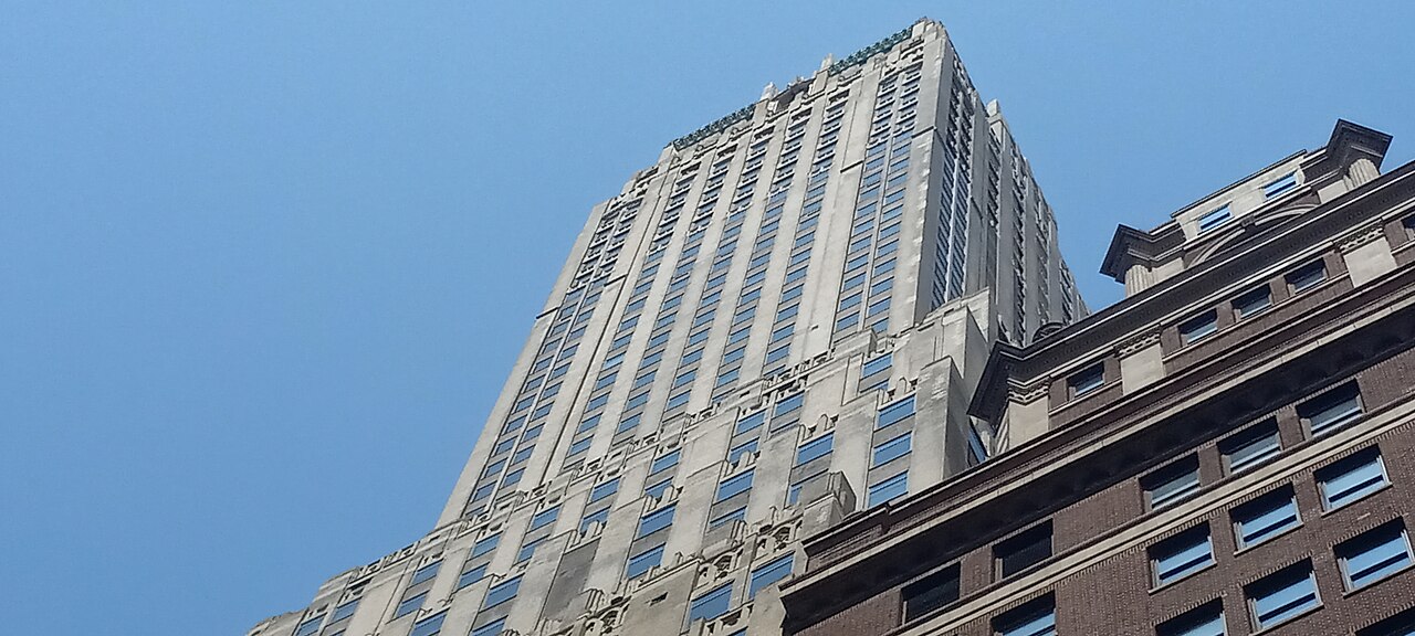 A pale stone skyscraper with stepped setbacks seen from street level against a clear blue sky.