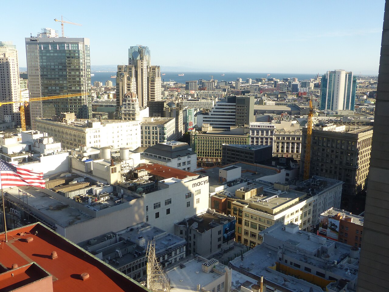 A high view over city rooftops and office towers stretching towards a hazy bay.