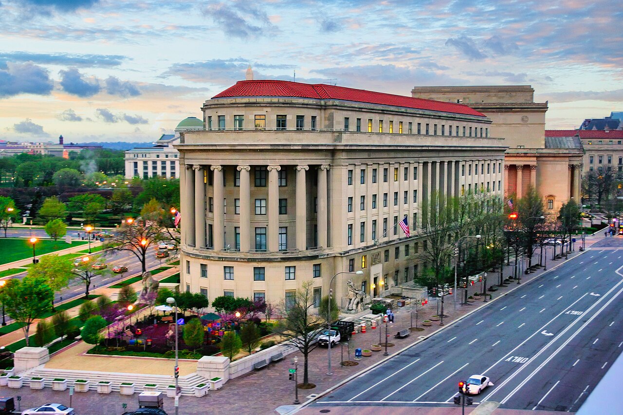 A columned stone building with a red tiled roof seen from above beside a wide avenue and flower beds.