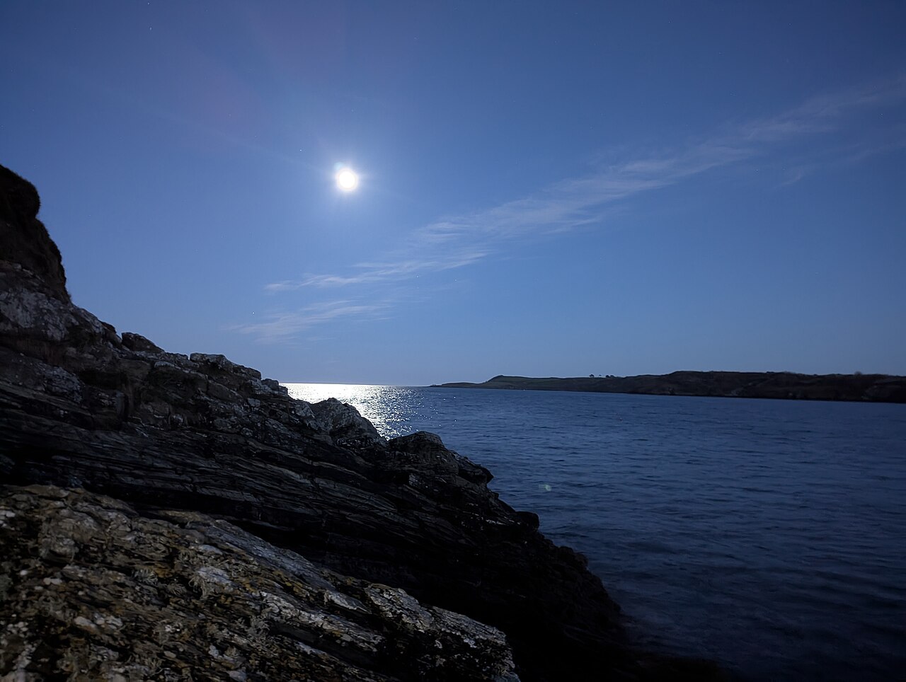 A bright moon hangs over dark water and a rocky shore, with a low headland on the horizon.