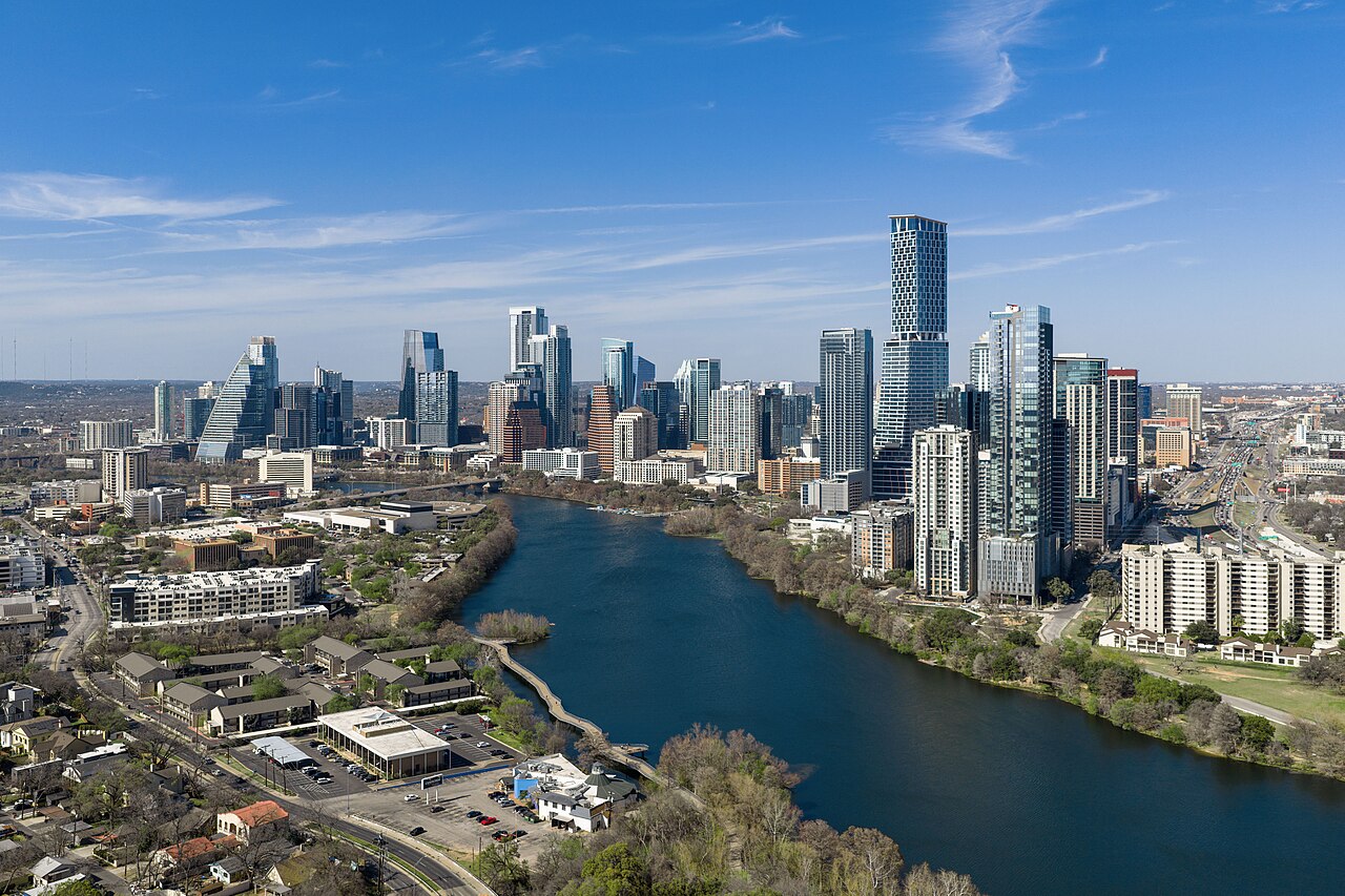 An aerial view of glass towers clustered beside a wide blue river that curves through a flat city.