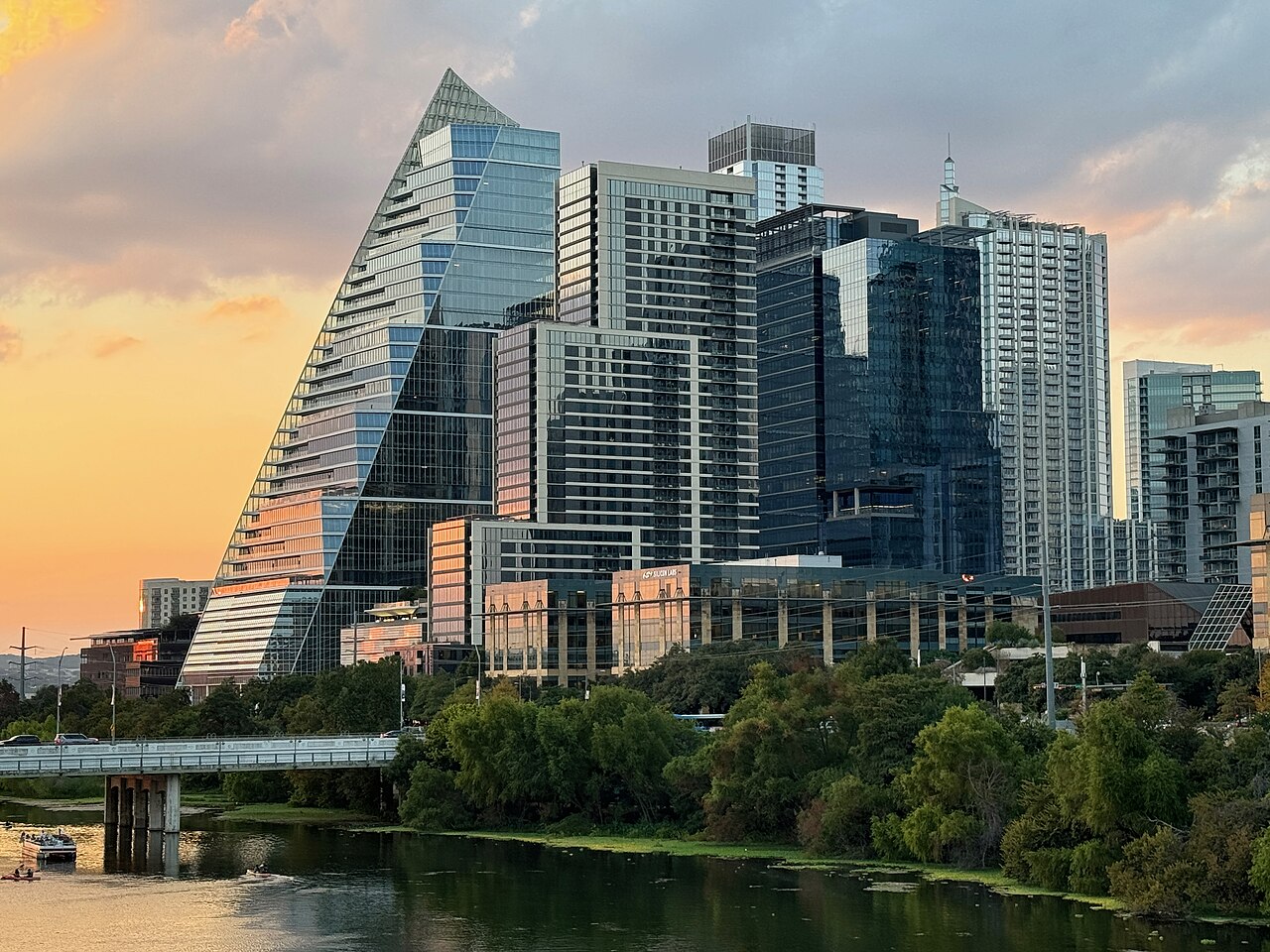 A sloping glass tower shaped like a sail stands among high-rises above a tree-lined lake at sunset.