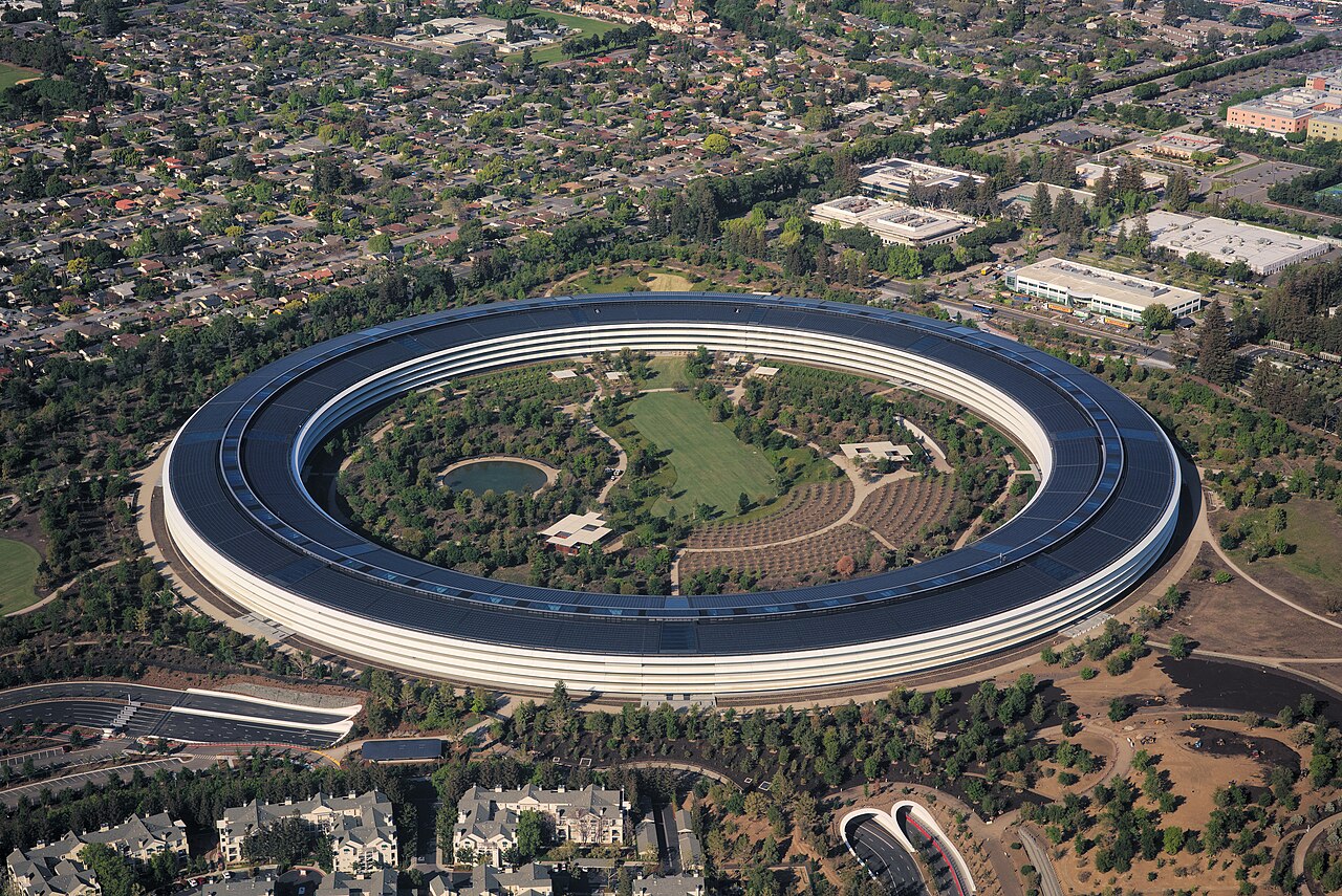 A huge ring-shaped building with a dark roof of solar panels encloses a green park, surrounded by suburbs.